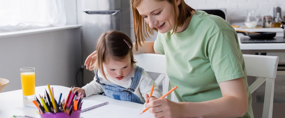 Parent and child with down syndrome drawing on paper near orange juice and fruits in kitchen